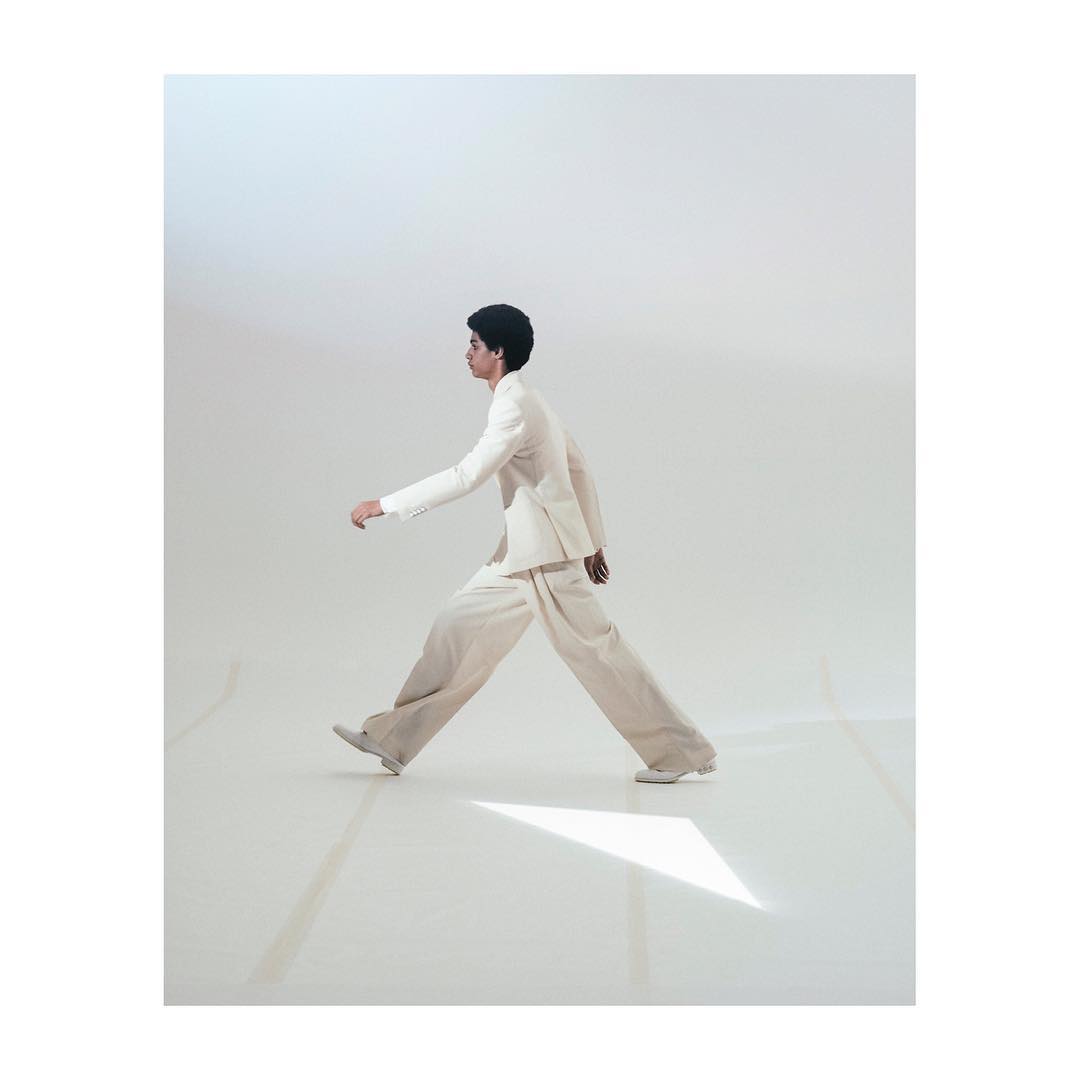 A slender adult strides purposefully across a stark white studio in a fashion editorial shot, wearing a white suit and exhibiting confidence.