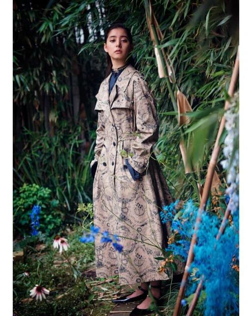 A young woman posing in a patterned trench coat, standing in a lush green garden with colorful flowers and bamboo stalks.