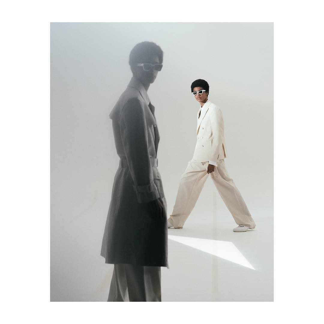 Two men in stylish suits and sunglasses strike poses in a bright studio setting. One wears a neutral coat, while the other sports a cream suit.
