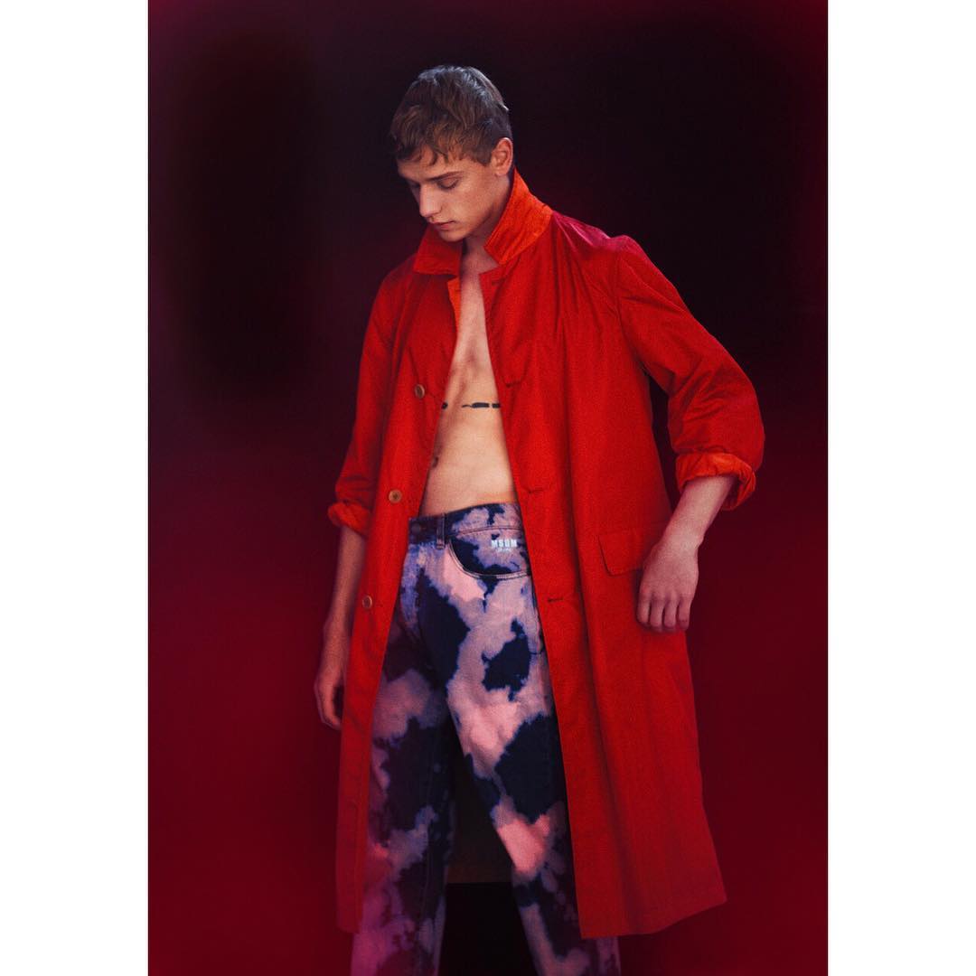 Young man in a red coat stands in front of a dark red background wearing patterned blue jeans, posing for a fashion portrait.