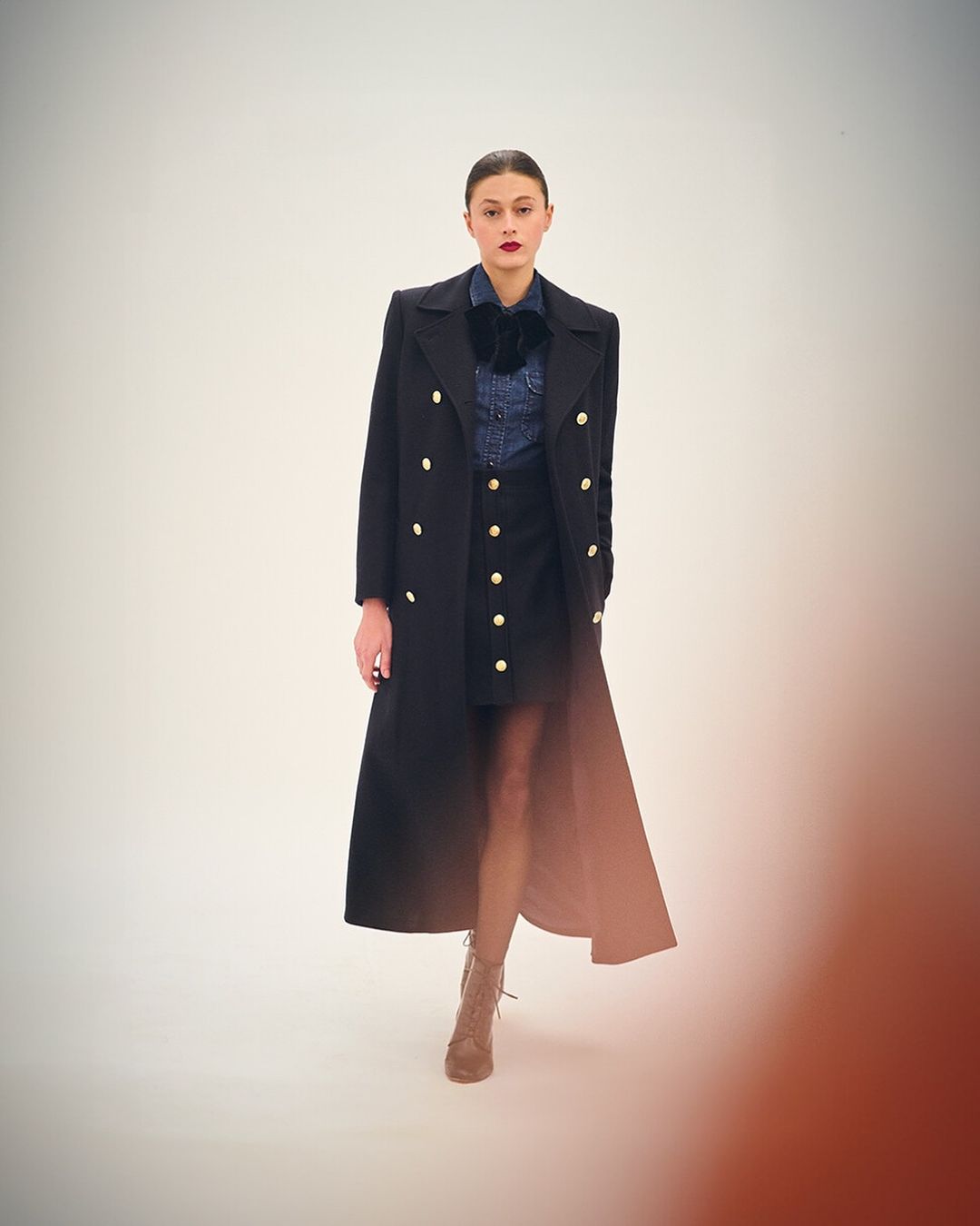 Model in long black double breasted coat with gold buttons over denim shirt and dark skirt by Serge Leblon