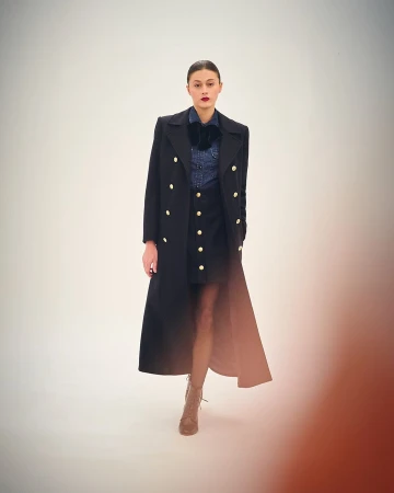 Model in long black double breasted coat with gold buttons over denim shirt and dark skirt by Serge Leblon