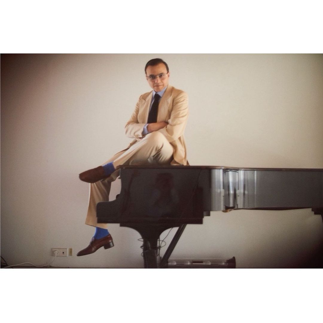 Man in beige suit and brown shoes poses on a grand piano with arms crossed in a bright studio setting.