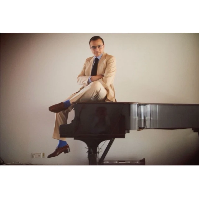 Man in beige suit and brown shoes poses on a grand piano with arms crossed in a bright studio setting.