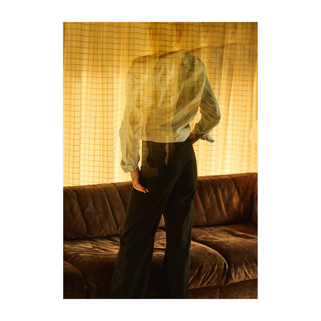 Back view of a woman in a white blouse and black trousers, posing indoors against a yellow curtain and brown couch in soft, diffused light.