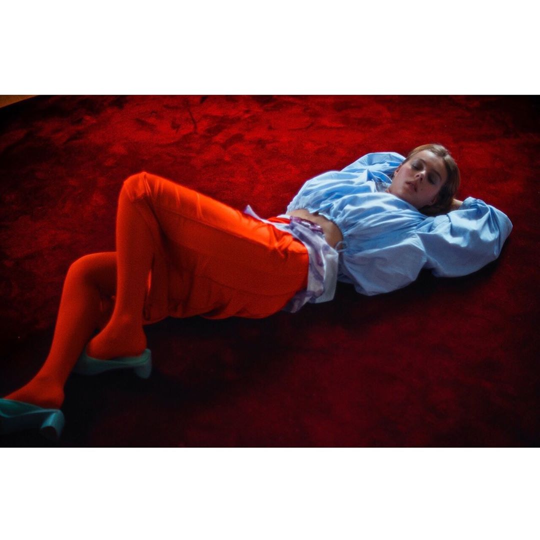 Fashion shot of a woman lying on a red carpet wearing a light blue blouse and orange pants, posing indoors with closed eyes.
