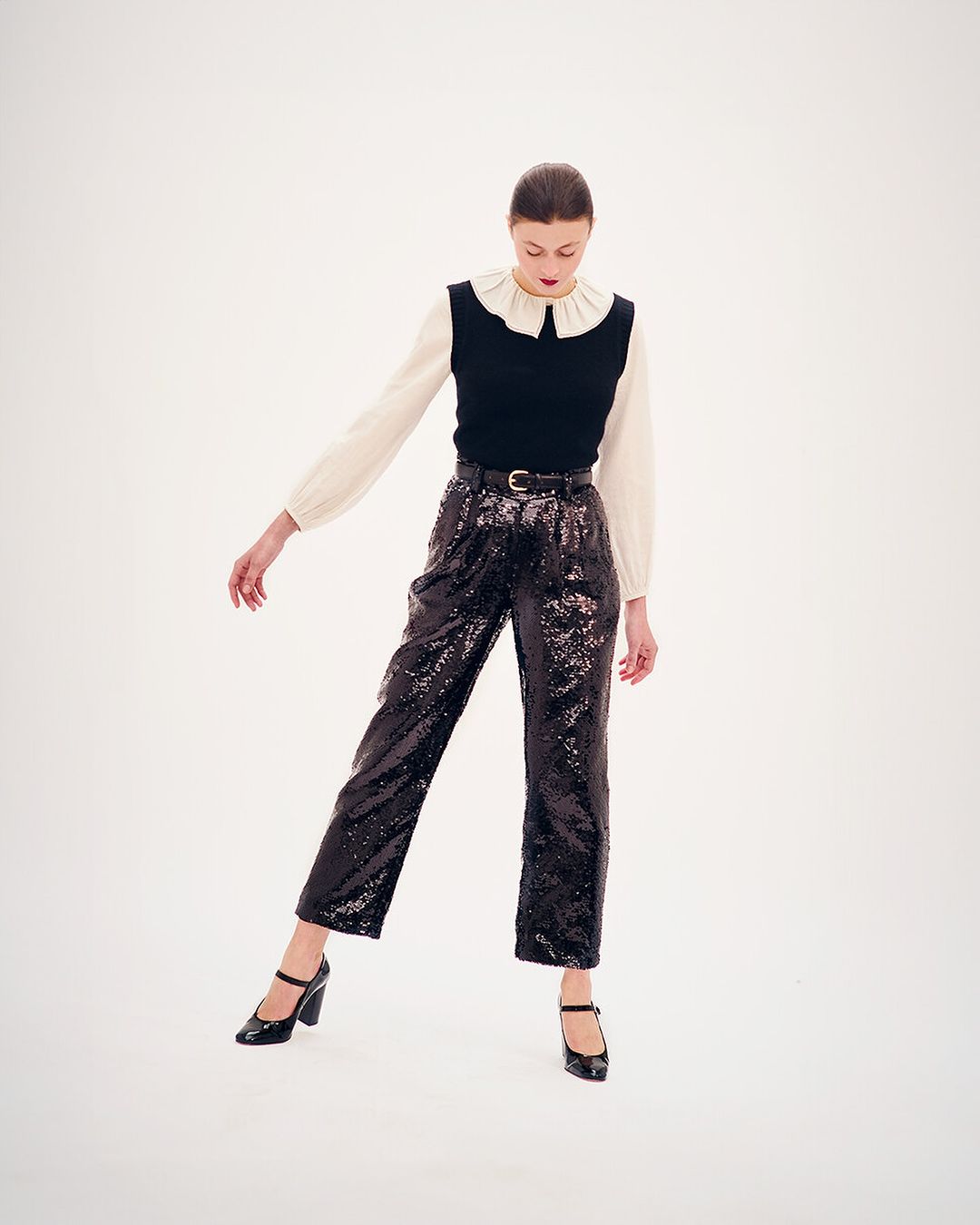 Elegant fashion shoot featuring a young woman in a white blouse, black vest, sequined pants, and Mary Jane shoes against a white backdrop.