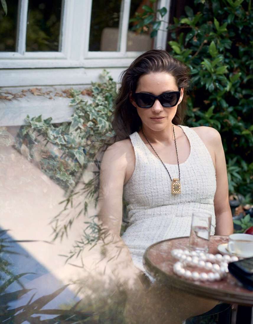 Portrait of a woman wearing black sunglasses and a white textured dress seated outdoors at a café table by Serge Leblon
