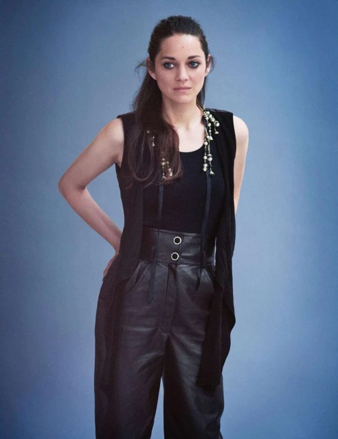 Portrait of Marion Cotillard in a black top and leather pants against a light blue background, with a long necklace detail.
