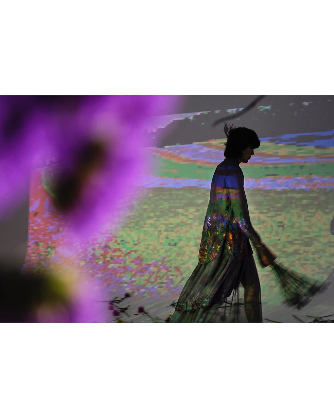 Fashion model walks on stage with purple floral foreground and background projection, creating an ethereal and artistic presentation.