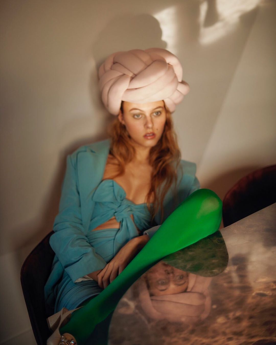 Portrait of a fair-skinned young woman with red hair wearing a blue dress, green leggings, and a pink head covering in a studio.