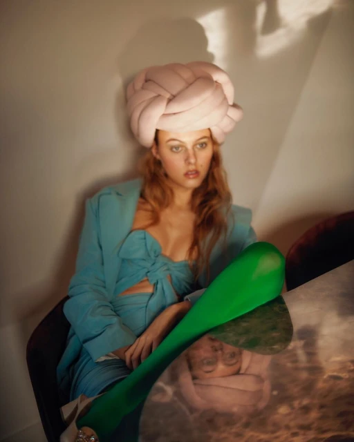 Portrait of a fair-skinned young woman with red hair wearing a blue dress, green leggings, and a pink head covering in a studio.