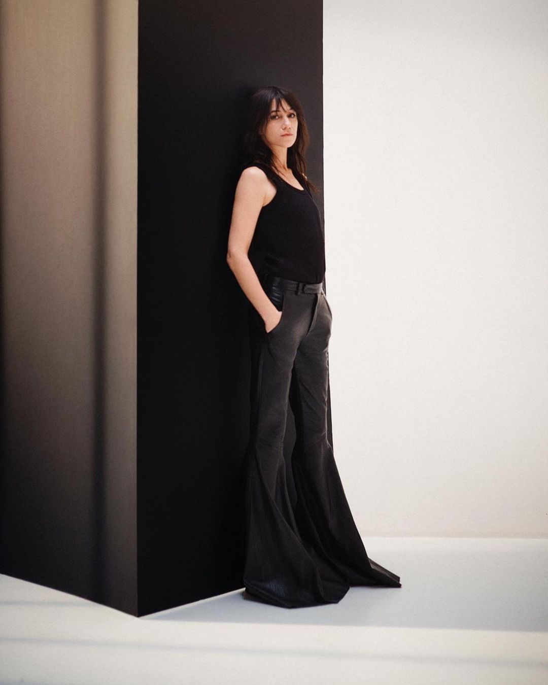 Studio shot of a woman with dark hair in a black top and pants, leaning against a black wall against a white wall backdrop. Fashion shoot.