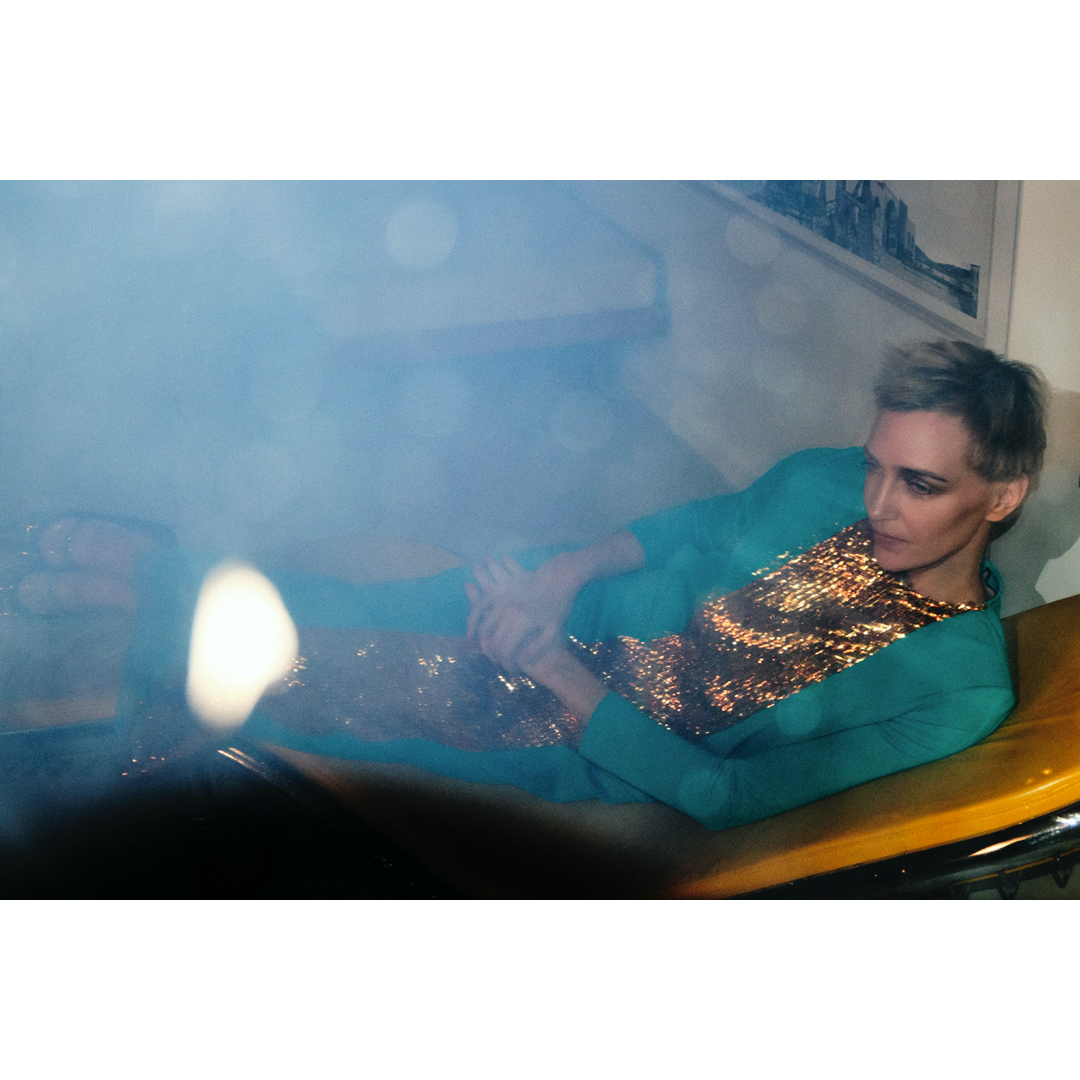 Woman in sequin dress and teal blazer reclines on a yellow surface in a hazy room with an abstract painting in background.