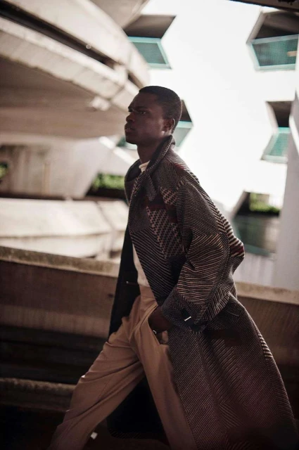 Stylish man in patterned coat walks in an urban setting with geometric concrete architecture, wearing tan pants, captured in medium shot.