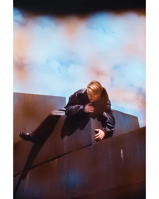 An adult male in a black leather jacket and black pants sits on a geometric concrete structure against a backdrop of a cloudy sky.