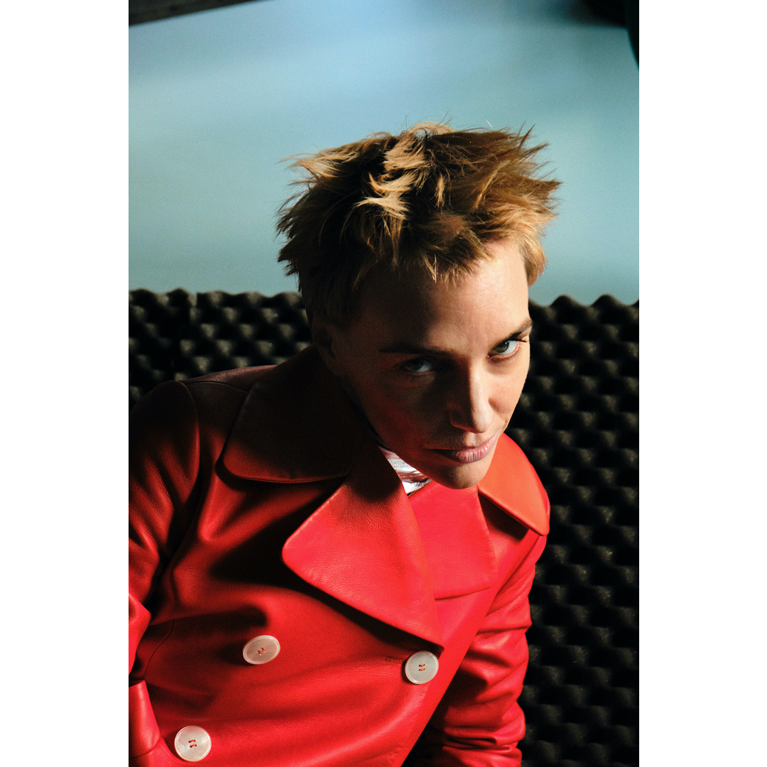 Portrait of a fair-skinned adult woman with short blonde hair, wearing a red pea coat with white buttons in front of a dark background.