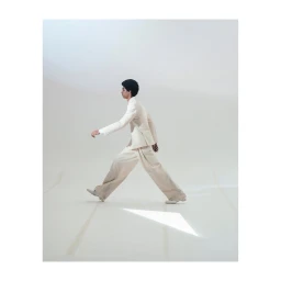 A man in a cream suit strides across a white studio floor in a side profile view