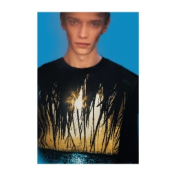 Portrait of a young male model wearing a black top with abstract gold graphic design against a blue gradient background.