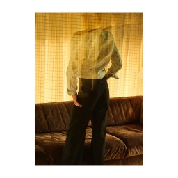 Portrait of a person standing in front of a tufted brown couch against a sheer grid curtain shot by Serge Leblon in Paris