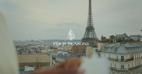 Eiffel Tower view with cityscape rooftops, sky, and Four Seasons logo. Urban Paris scene with landmark architecture.