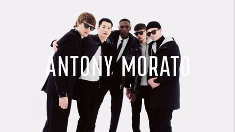 Five male models in black coats and sunglasses posing together against a plain white background with bold white text overlay.