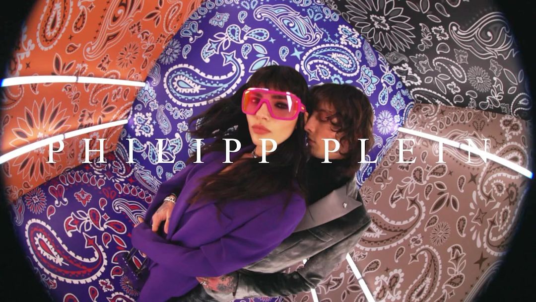 Fashion shot of a woman in pink sunglasses and a man embracing amidst a backdrop of colorful patterned fabrics and neon lighting.