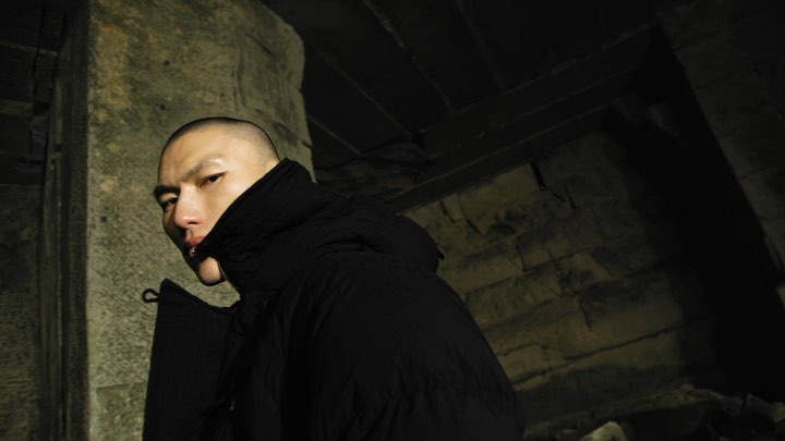 Portrait of a person in a black puffer jacket looking over their shoulder, photographed against a weathered stone wall.