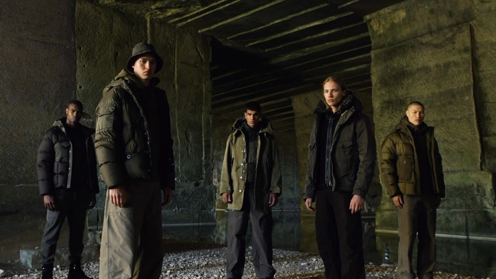 Five people in hooded jackets pose indoors against a stone wall, showcasing a fashion collection with dark neutral tones and relaxed style.