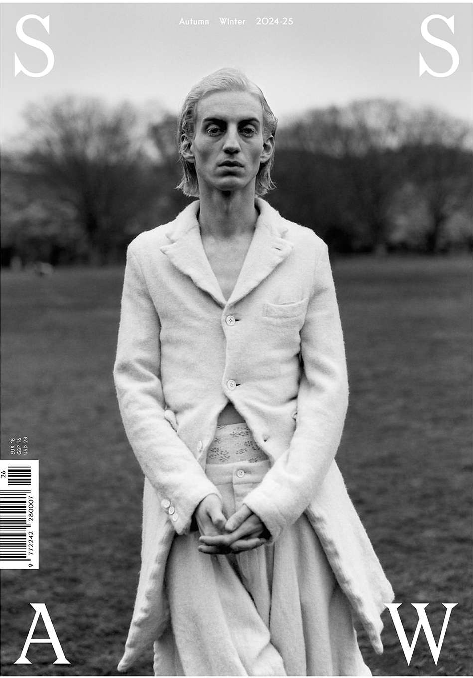 Black and white magazine cover shot of a thin adult male in a white suit standing in a grassy field with trees in the background.