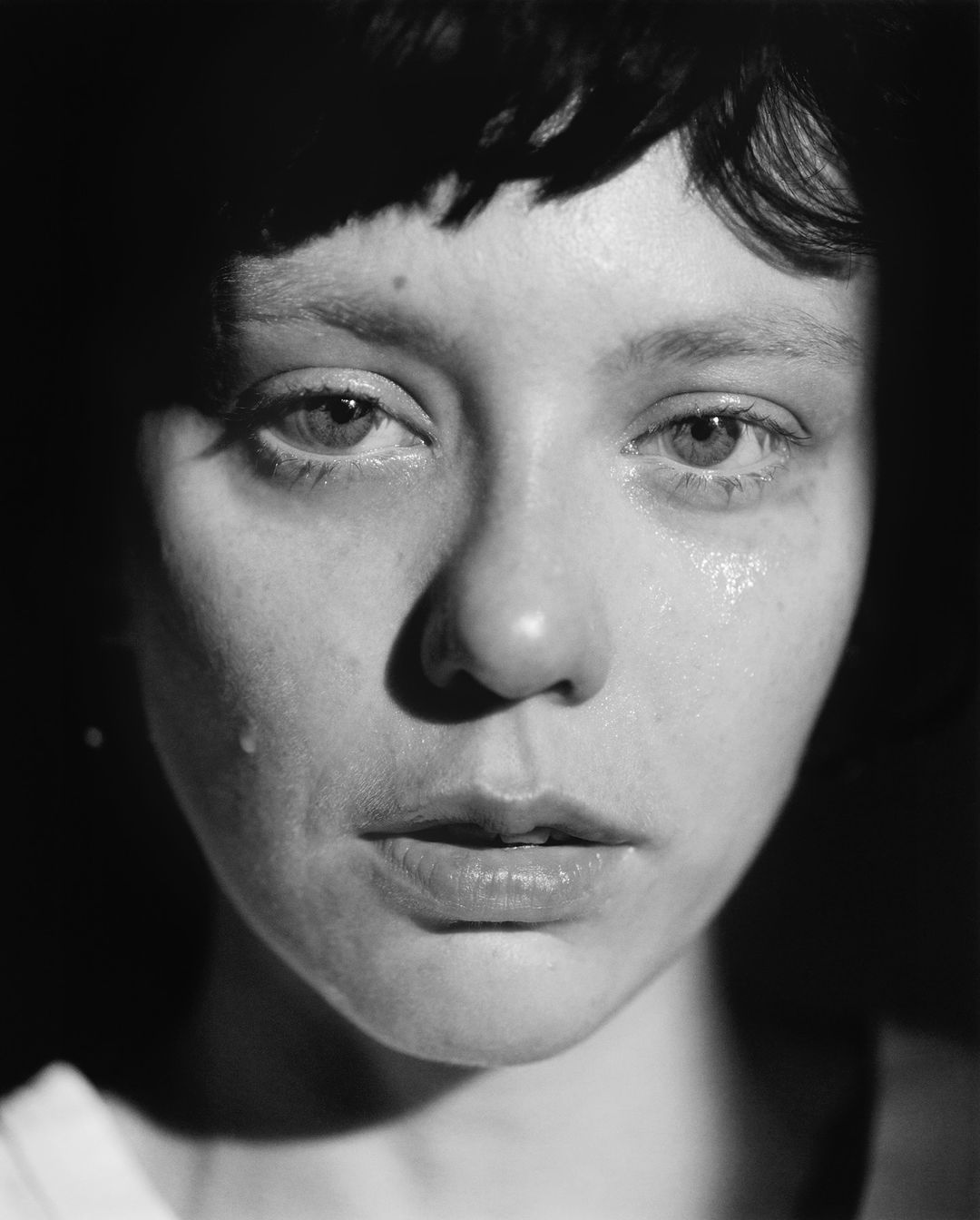 Close-up monochrome portrait of a young adult woman with a somber expression, visible tears, and dark, short hair, set against a dark background.
