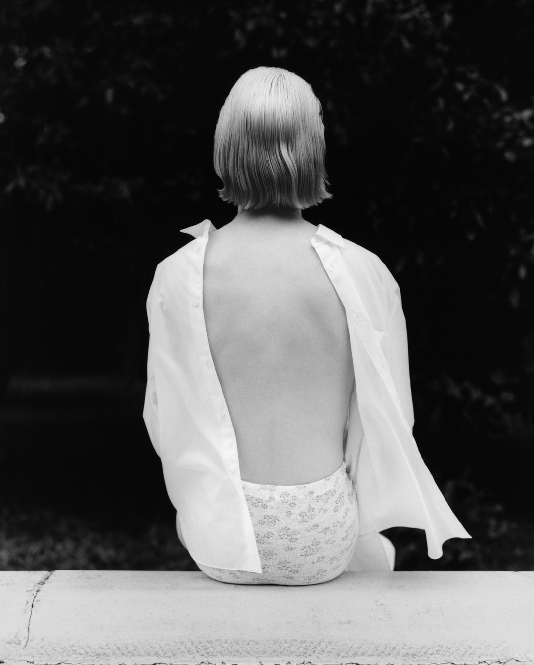 Monochrome portrait of a person from behind with open white shirt, sitting outdoors against a dark foliage background.