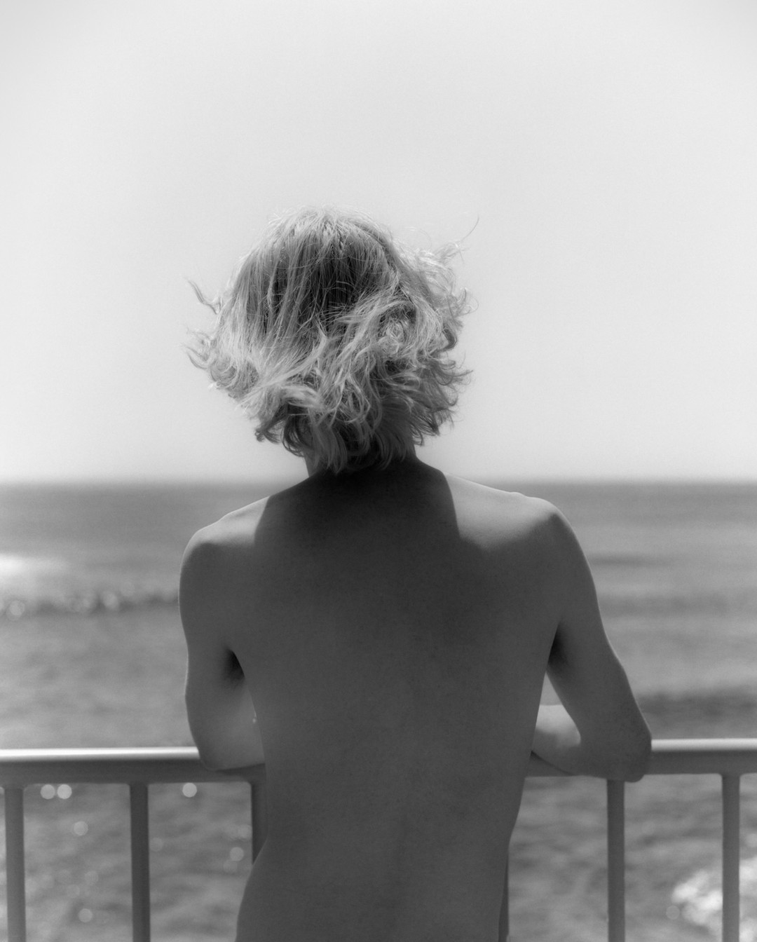 Monochrome shot of an adult leaning on a railing, facing the ocean on a bright day. The person's back and curly hair are visible.