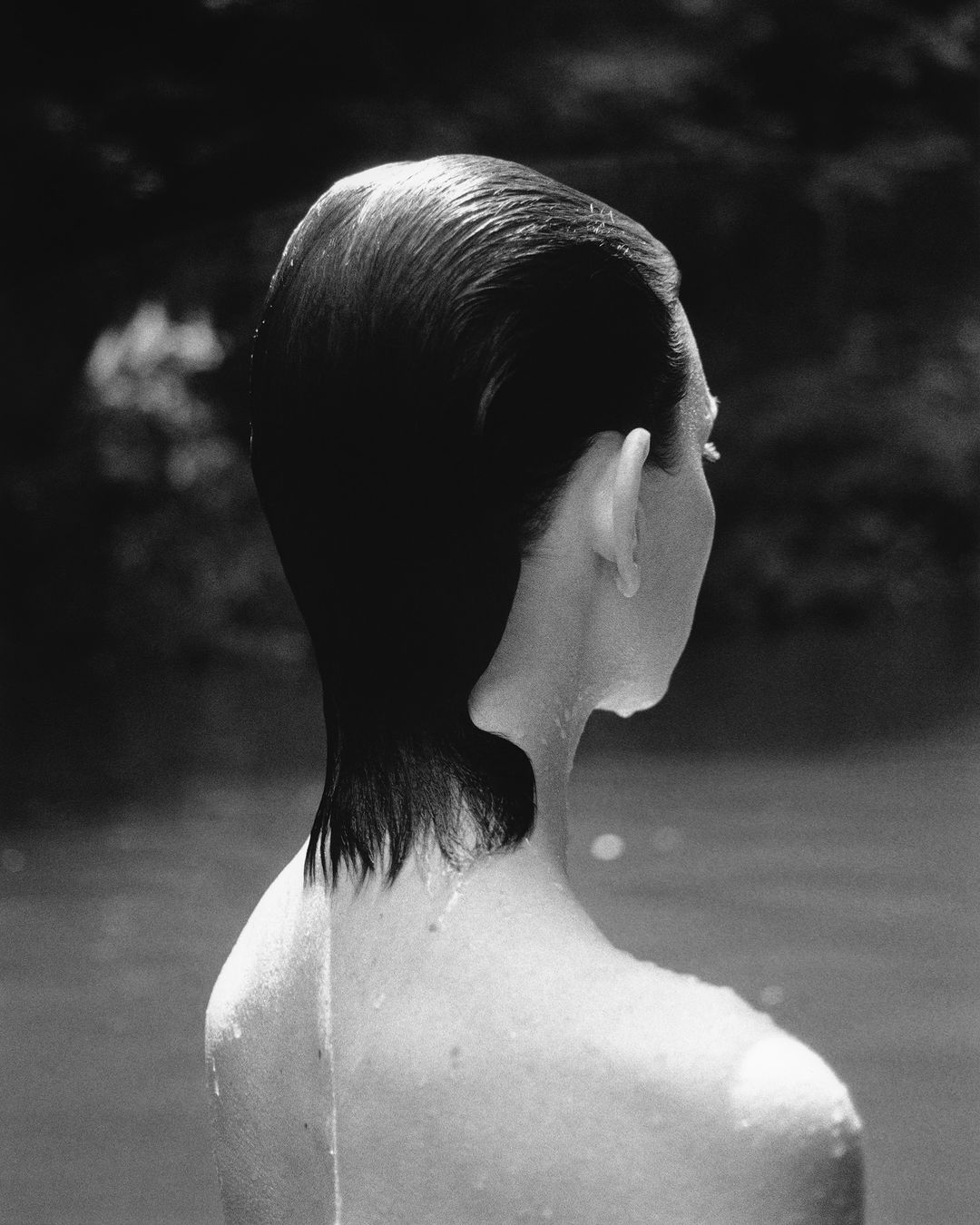 Back view of a person with wet, slicked-back hair standing in the water, captured in monochrome. The background is blurred and indistinct.