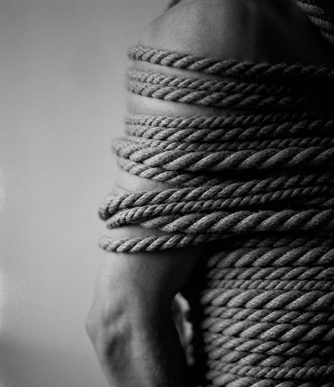 Close-up in black and white of an adult torso and arm, wrapped tightly with natural rope against a blurred grey background, studio lighting.