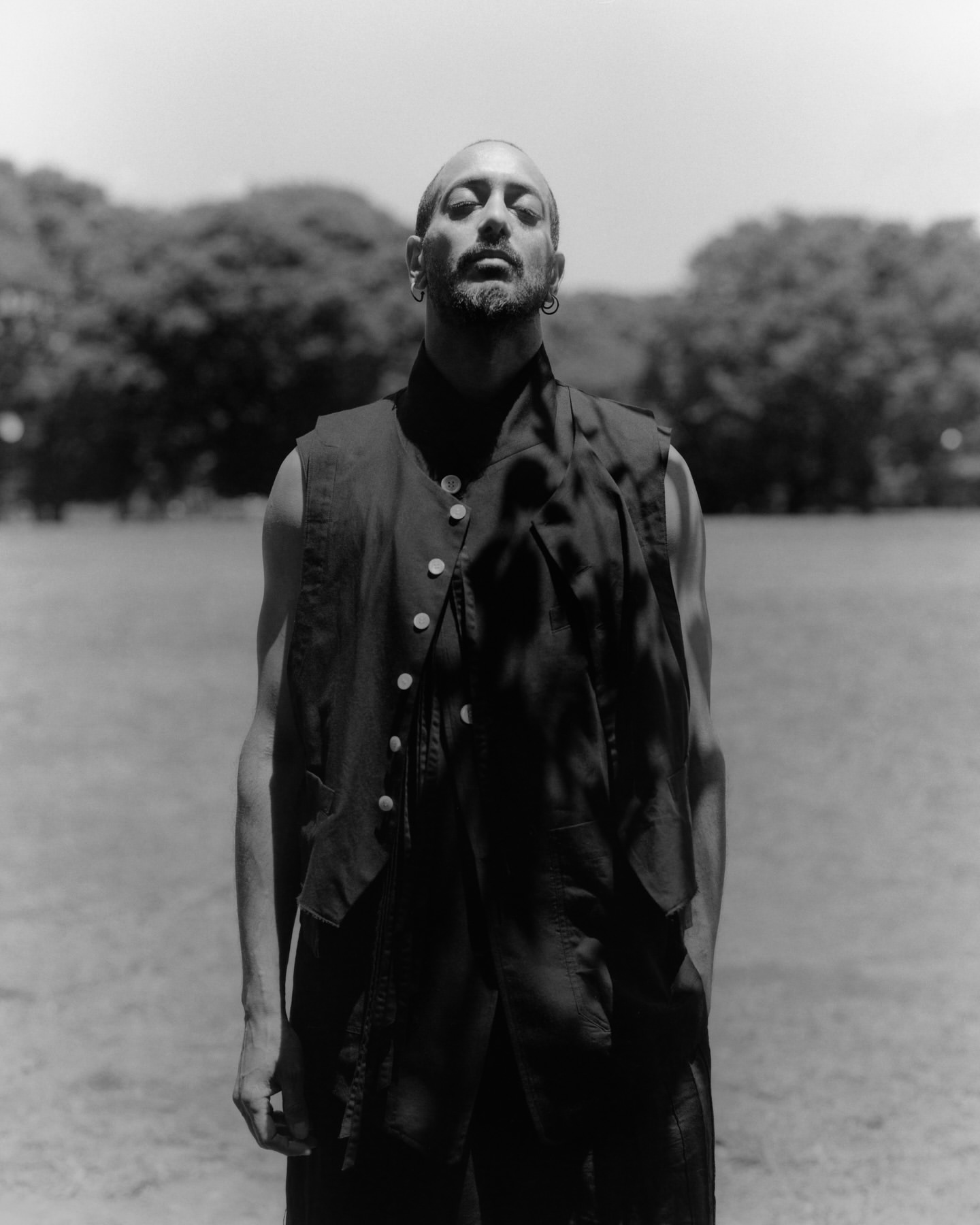 Monochrome portrait of adult male outdoors in vest. Neutral expression, grassy field and trees in background.