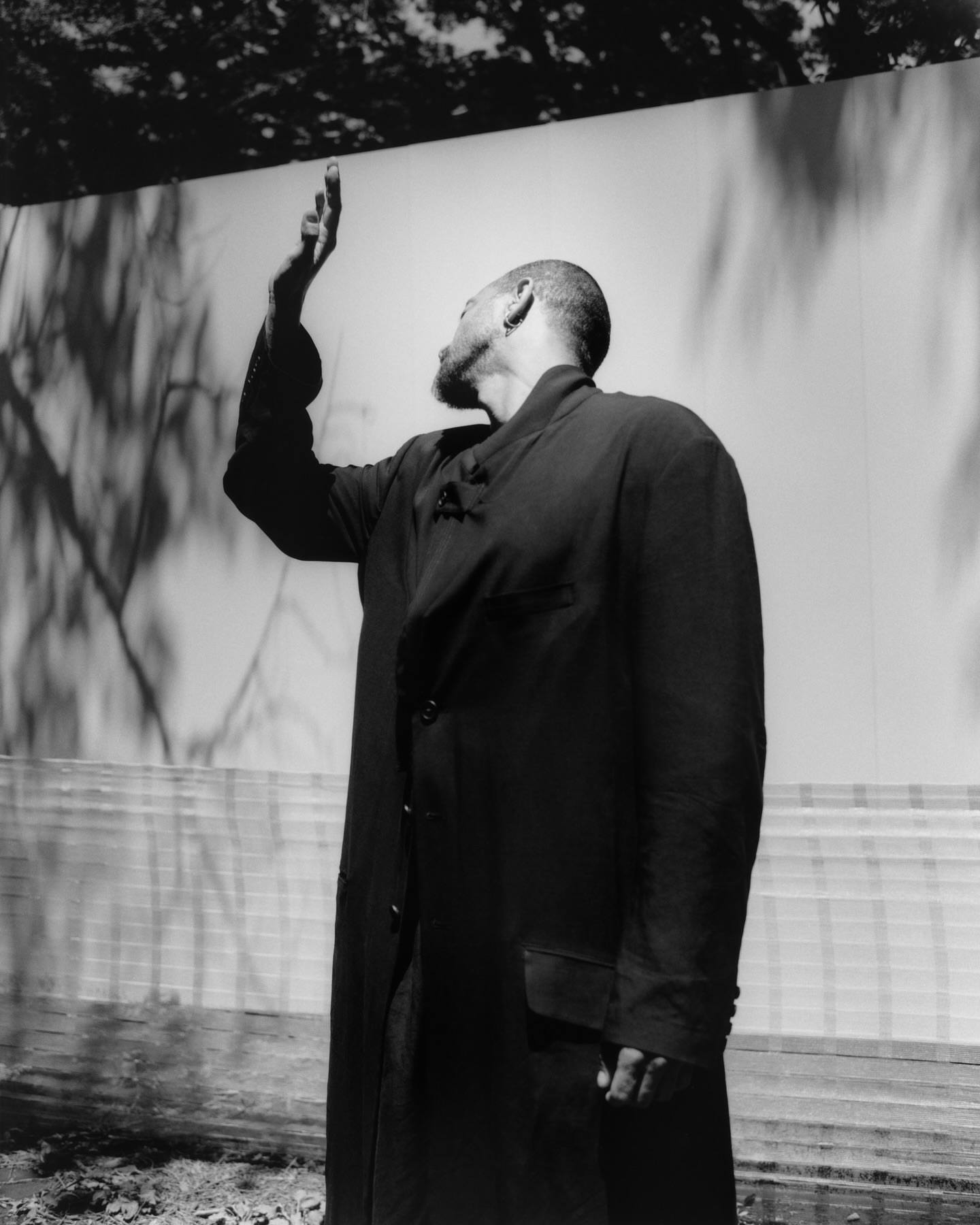 Monochromatic portrait of a Black adult male in a long coat with a raised arm, standing in front of a bright wall with tree shadows.