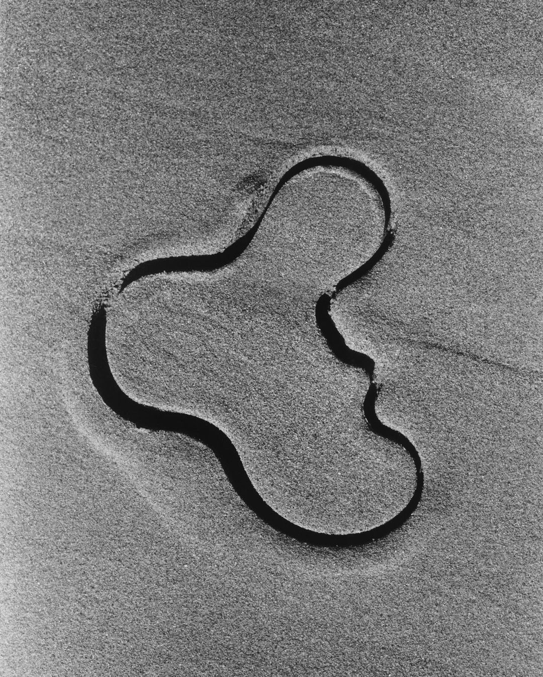 Black-and-white photograph of an abstract, fluid shape etched into the sand on a beach, creating a textured pattern.
