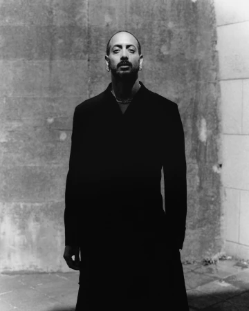 Portrait of a man in a black jacket standing against a textured concrete wall, photographed in monochrome.