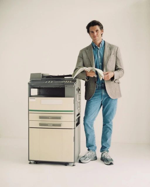 A man in a blazer and jeans stands next to a copy machine, holding a stack of papers, indoors with a neutral background.