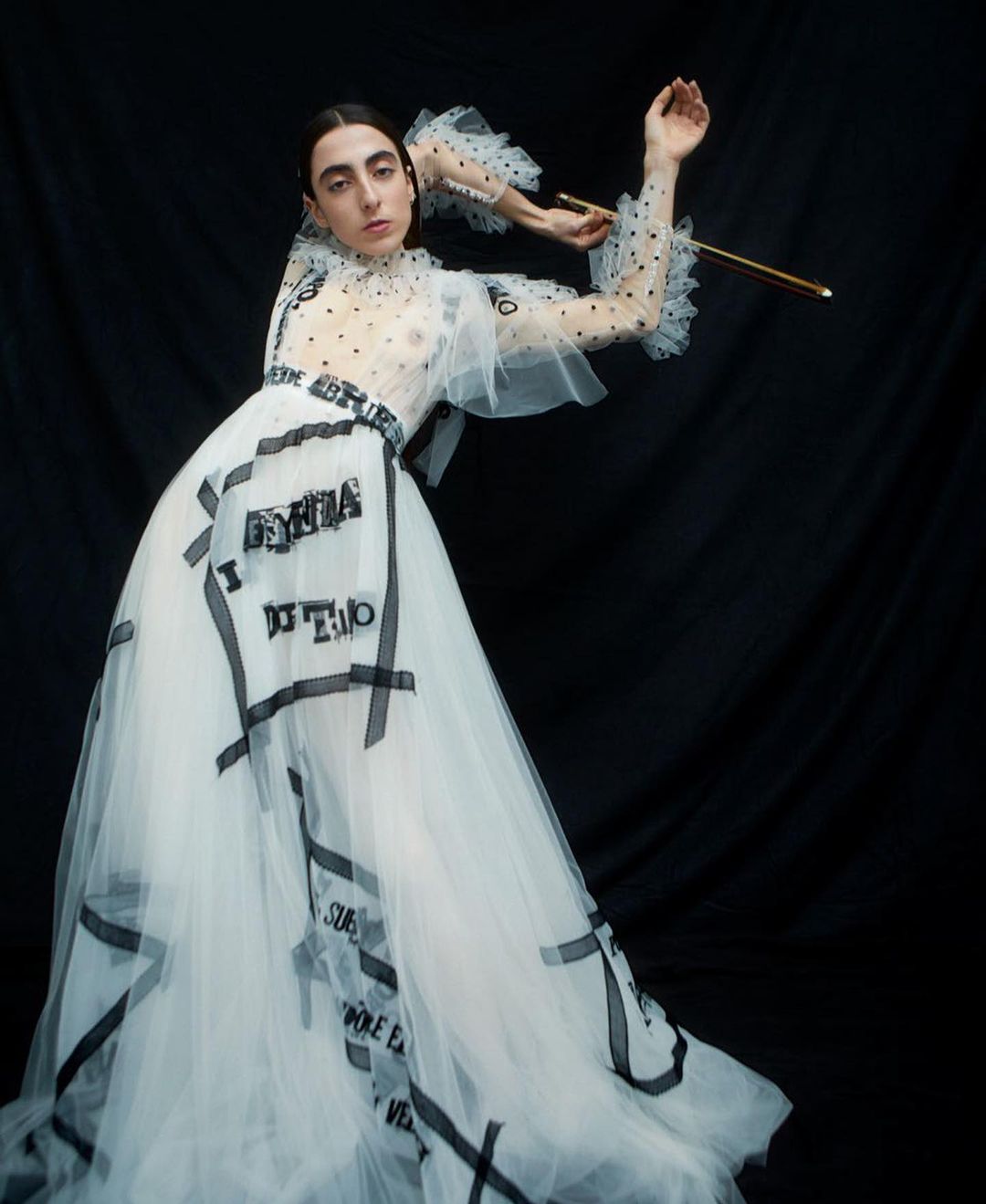 Woman poses in a white, tulle gown with black text detailing, accentuated by a black backdrop. Her makeup is natural, and she's holding a golden object.