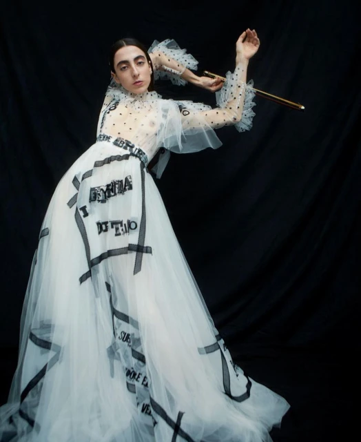 Woman poses in a white, tulle gown with black text detailing, accentuated by a black backdrop. Her makeup is natural, and she's holding a golden object.