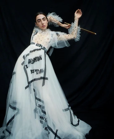 Woman poses in a white, tulle gown with black text detailing, accentuated by a black backdrop. Her makeup is natural, and she's holding a golden object.