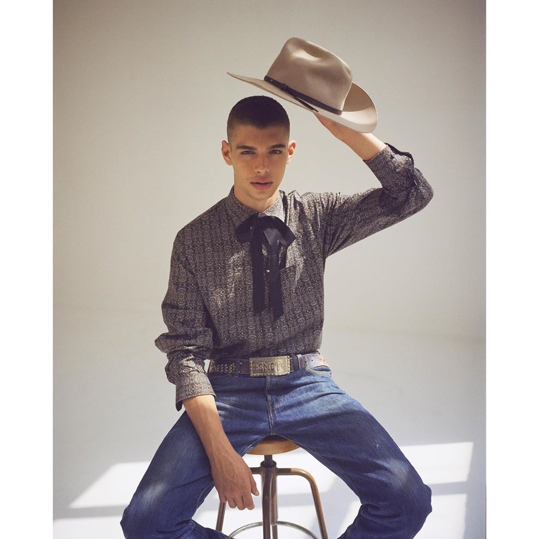 A young adult male sits on a stool indoors, holding a beige cowboy hat aloft. He wears a patterned shirt and jeans in soft, neutral lighting.