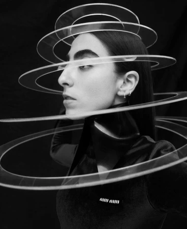 Monochrome portrait of a woman in a dark top, wearing a transparent ring headpiece, with a blurred black background, fashion-editorial style.