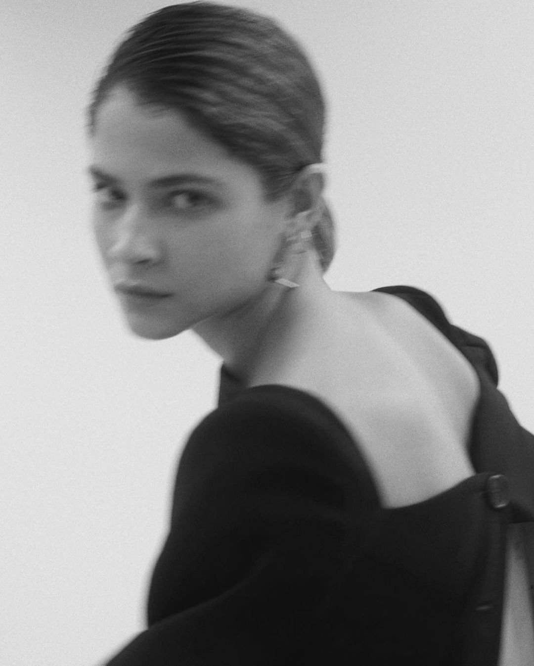 Black-and-white portrait of a young adult female in a dark dress, looking over her shoulder with slicked-back hair against a bright background.