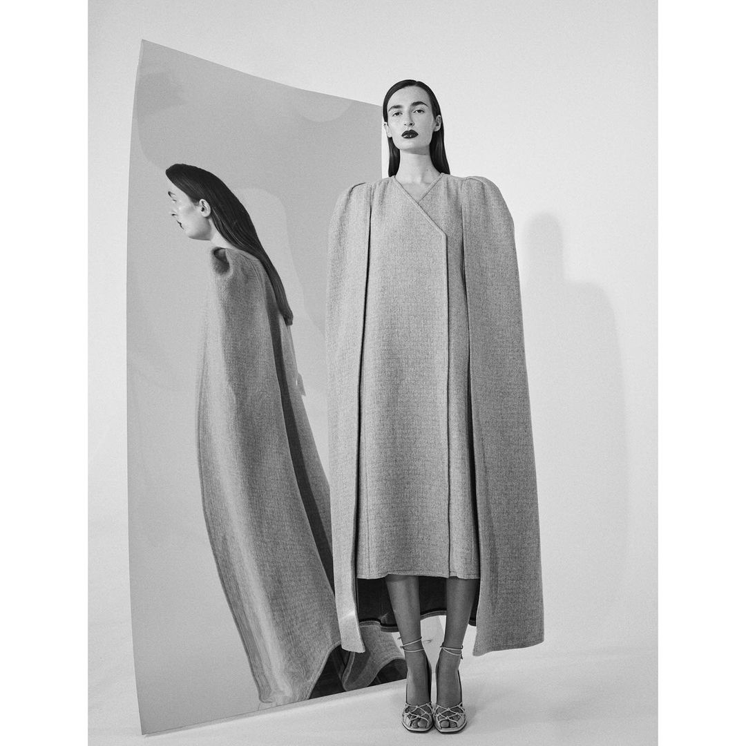 Monochrome portrait of a woman in a long grey coat with shoulder pads, standing in front of a reflective screen.