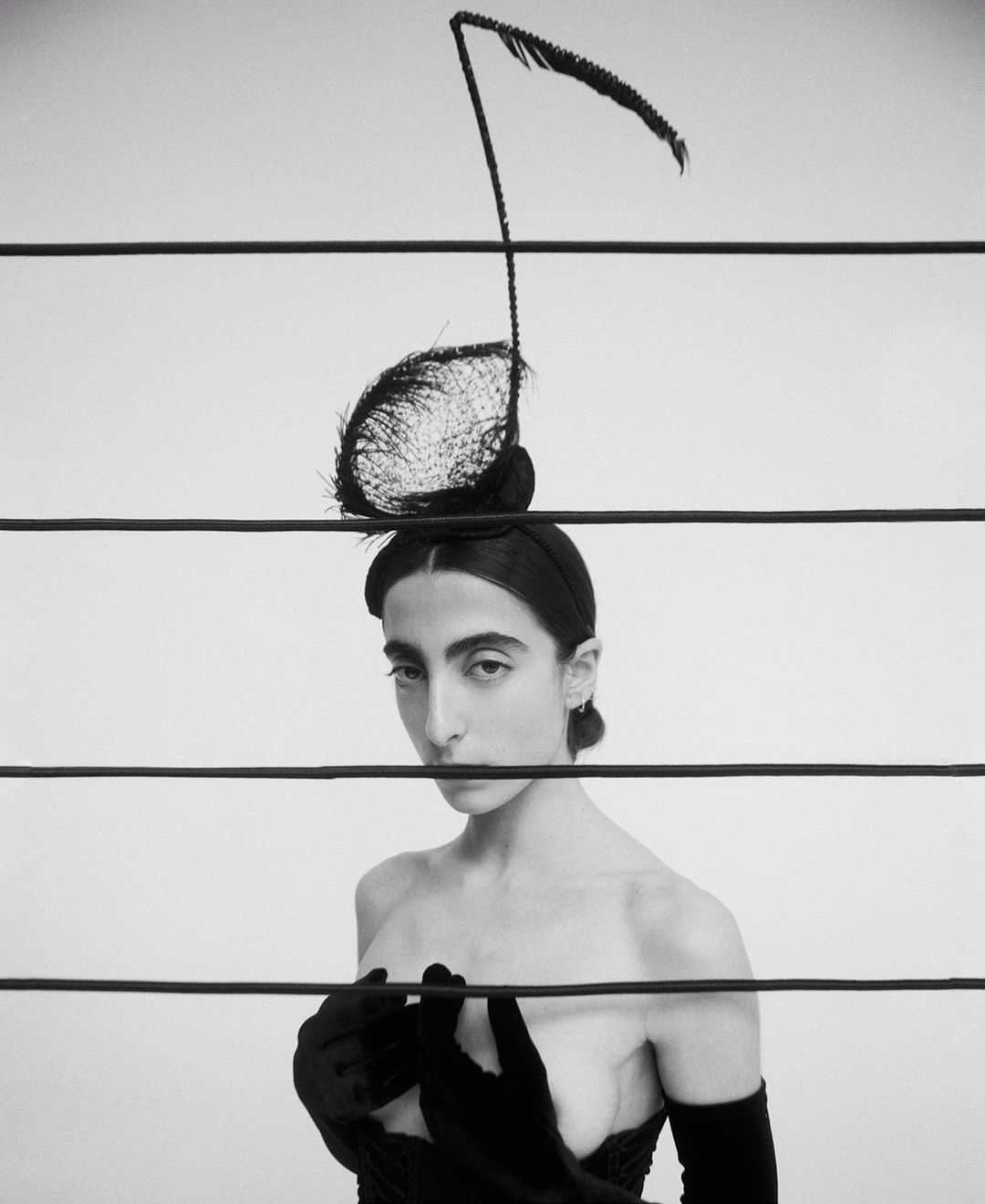Striking black-and-white portrait of a woman with a distinctive feathered headpiece, gazing through parallel black lines in a studio setting.