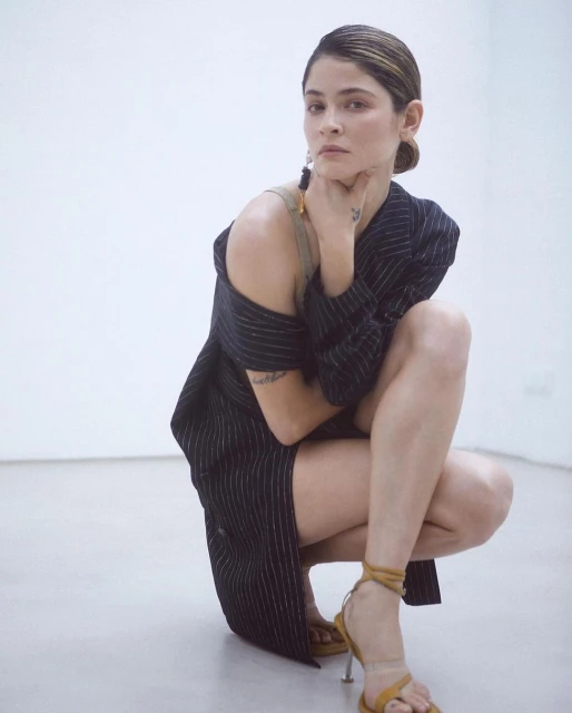 Young woman poses in a black and white pinstripe blazer and tan heels in front of a white wall, seated in a squatting position.