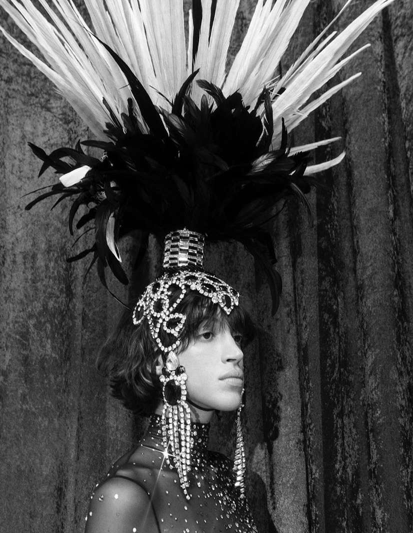 Striking portrait of a person wearing an elaborate feather headdress and costume with crystal embellishments, set against a draped backdrop.